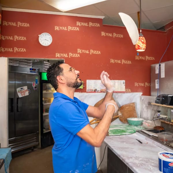 A Royal Pizza chef in a blue shirt expertly tosses pizza dough in the air.