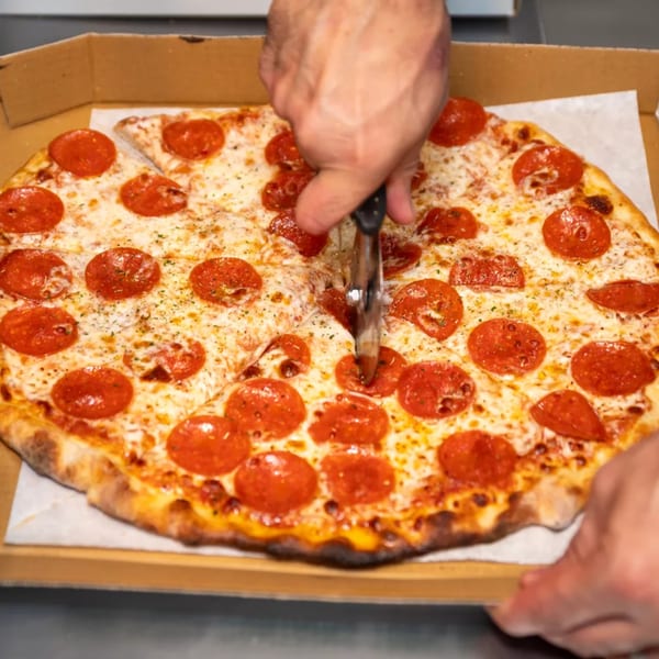 Hands cutting a Royal Pizza pepperoni pizza in a cardboard box with a pizza cutter.