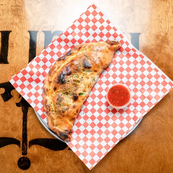 A golden-brown calzone with herbs and a side of red sauce on checkered paper at Royal Pizza.