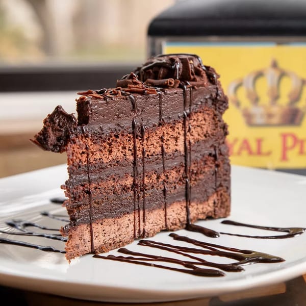 A rich slice of Royal Pizza's chocolate cake with chocolate frosting and drizzle on a white plate.