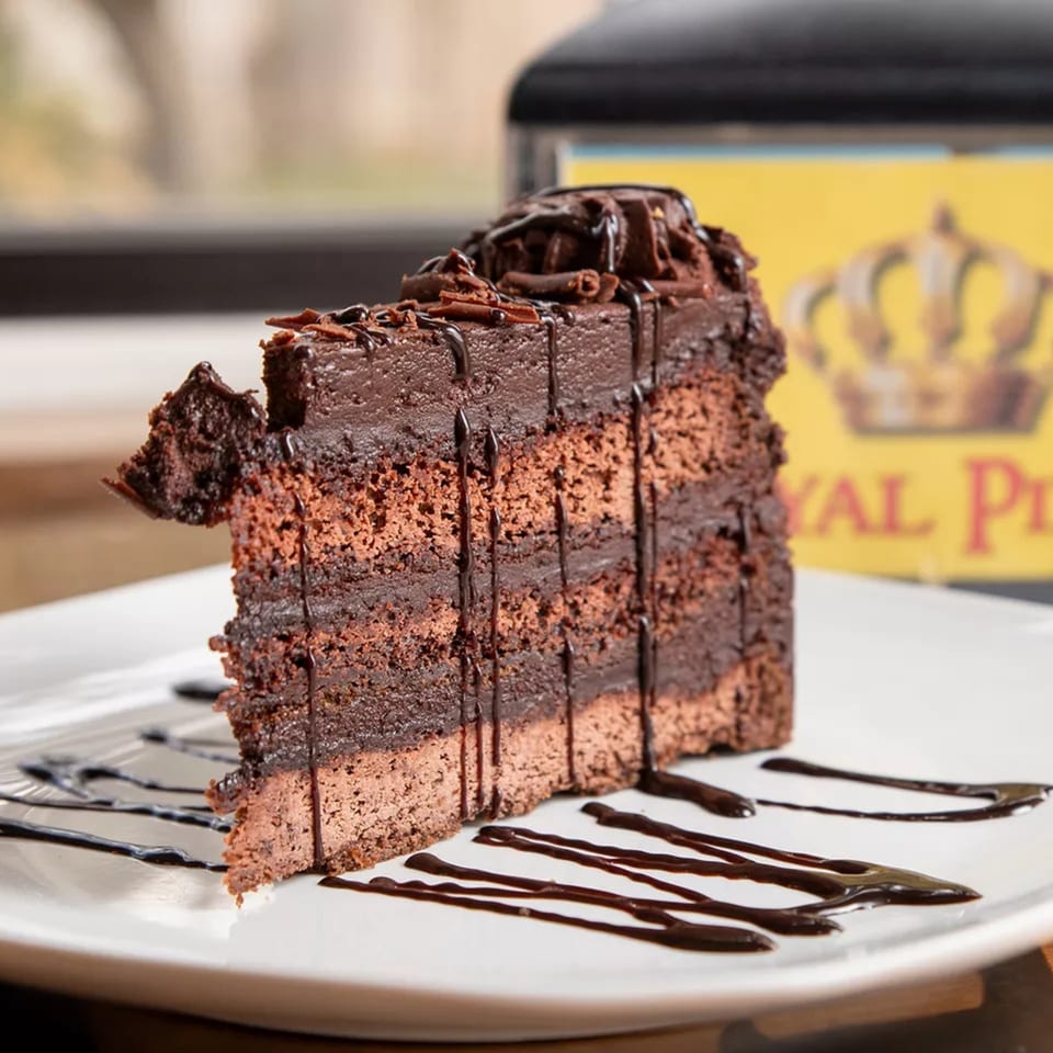 A rich slice of Royal Pizza's chocolate cake with chocolate frosting and drizzle on a white plate.