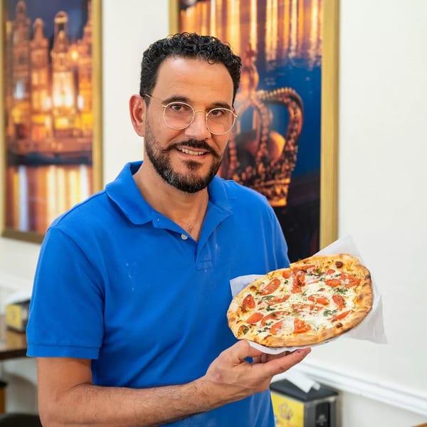 A smiling man in a blue polo shirt holds a freshly baked Royal Pizza Margherita pizza.