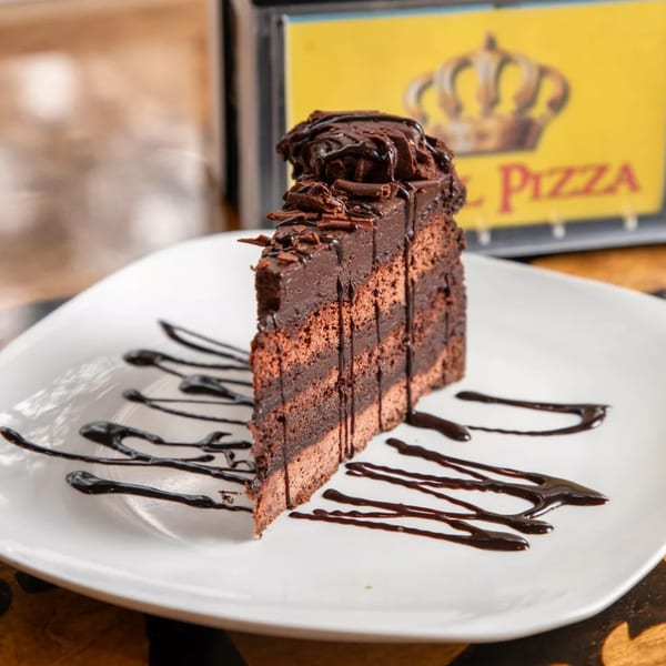 A slice of Royal Pizza's rich chocolate cake with chocolate shavings and drizzle on a white plate.