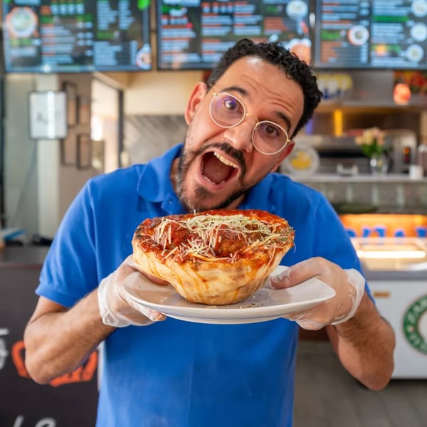 Man in glasses and blue shirt holding a deep-dish pizza with meatballs and cheese, mouth open.