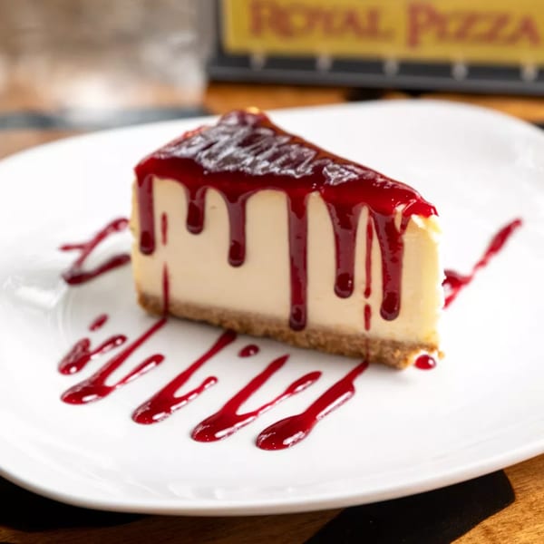 A slice of cheesecake with raspberry drizzle on a white plate, with a Royal Pizza sign in the background.