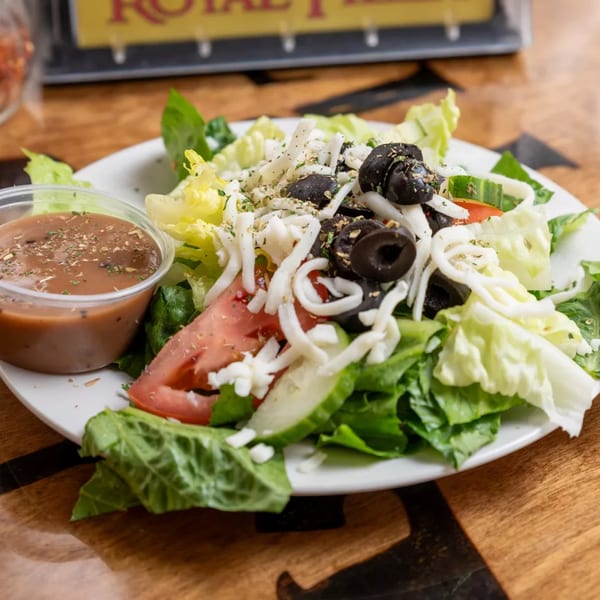 A Royal Pizza side salad with lettuce, tomatoes, cucumbers, black olives, shredded cheese, and dressing.