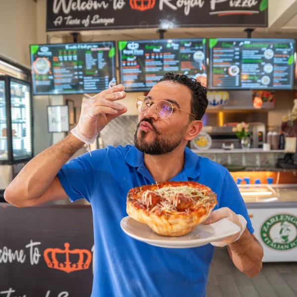 A Royal Pizza chef in a blue shirt holds a deep-dish pizza, smelling it with delight.